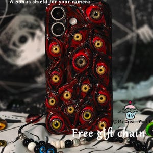 May include: A red and black phone case with a pattern of multiple eyes, with yellow irises and brown pupils. The case is for iPhone 13 series and above models. A black and white beaded necklace is also visible.