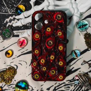 May include: A red and black phone case featuring a pattern of numerous yellow-eyed, bloodshot eyes. The case has a glossy finish and a textured, organic design. The case is surrounded by various decorative eye-shaped cabochons.