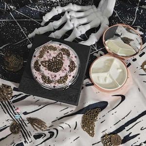 May include: A round, pink compact mirror with a decorative butterfly design, rhinestones, and ornate metal accents. The mirror is open, revealing a compartment with three sections. A black box and decorative metal pieces surround the mirror.