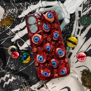 以下が含まれることがあります： 青い虹彩を持つ複数のリアルな眼球で覆われた赤い携帯電話ケース。 ケースは、スケルトンハンド、ピン、装飾的な目の形をしたオブジェクトなど、他の不気味なアクセサリーに囲まれています。