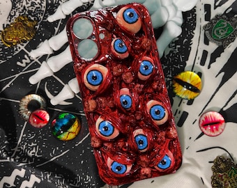 Puede incluir: Una funda de teléfono roja cubierta con múltiples globos oculares de aspecto realista con iris azules. La funda está rodeada de otros accesorios espeluznantes, incluyendo una mano de esqueleto, alfileres y objetos decorativos en forma de ojo.