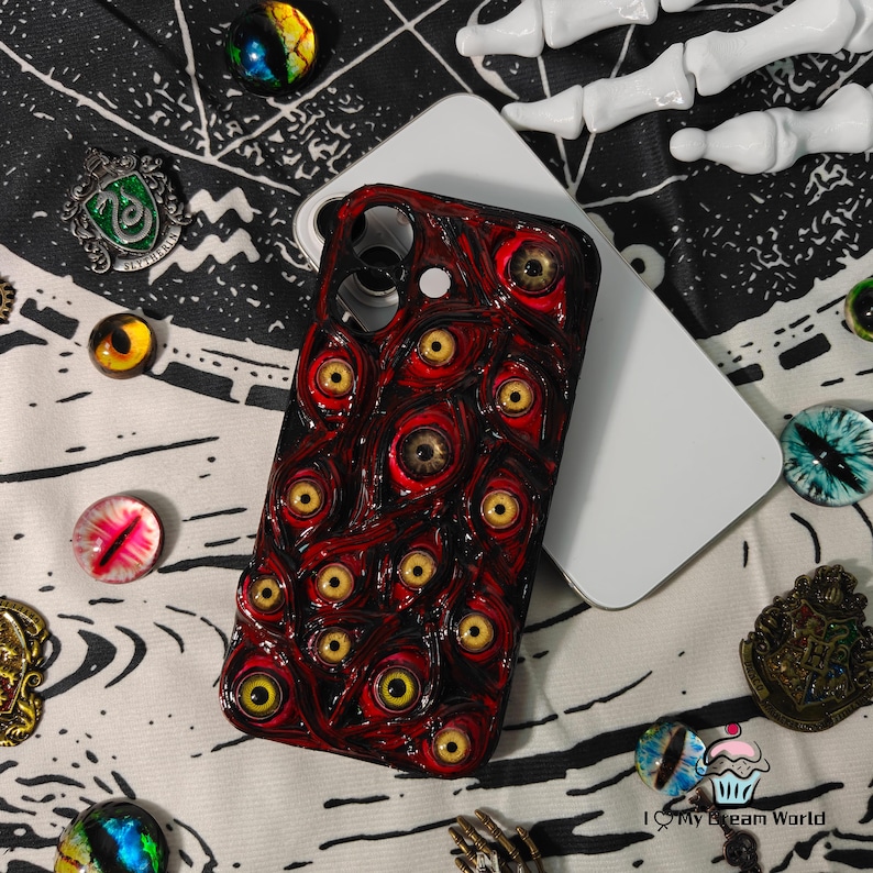 May include: A red and black phone case featuring a textured design of numerous yellow-eyed eyeballs. The case is resting on a white phone, surrounded by decorative glass eye cabochons and metal charms.