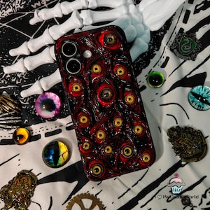 May include: A dark red phone case covered in multiple yellow-eyed, red-rimmed eye designs. The case is surrounded by various decorative items, including glass eyes, pins, and a white skeleton hand, creating a spooky, Halloween-themed aesthetic.