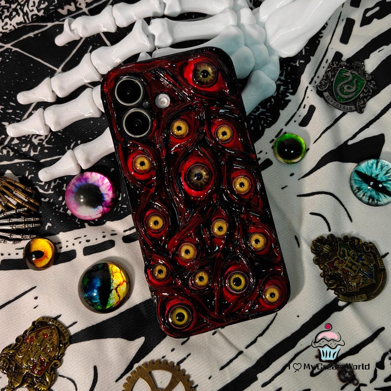 May include: A dark red phone case covered in multiple yellow-eyed, red-rimmed eye designs. The case is surrounded by various decorative items, including glass eyes, pins, and a white skeleton hand, creating a spooky, Halloween-themed aesthetic.