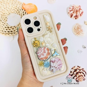 May include: A cream-colored phone case decorated with seashells, a gold sun charm, and a starfish. The case features a variety of shells in shades of white, pink, and orange, with a blue and gold tail charm. The case is designed for a smartphone.