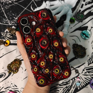 Könnte beinhalten: Eine Handyhülle mit einem dunkelroten und schwarzen Design mit mehreren augenförmigen Verzierungen mit gelben Iris. Die Hülle hat eine glänzende Oberfläche und wird in einer Hand gehalten. Ein einzigartiges, künstlerisches Accessoire.