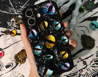 May include: Black phone case featuring multiple colorful, detailed eye designs. The eyes have various colors, including blue, yellow, and green, set against a textured black background. The case is designed for a smartphone and has camera cutouts.