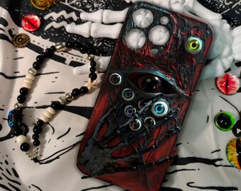Puede incluir: Una funda de teléfono roja y negra con múltiples diseños de ojos. La funda tiene una apariencia texturizada y presenta un ojo central grande y varios ojos más pequeños. También se ve un brazalete de cuentas negras y otros elementos decorativos.