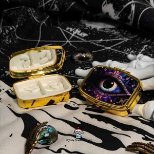 May include: Two gold-colored pill boxes with an open and closed lid. The open box contains pills and a mirror. The closed box features an eye design with purple and blue accents. The boxes are on a black and white patterned surface.
