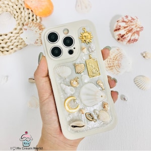 May include: Cream-coloured phone case adorned with seashells, pearls, and gold charms, including a sun, pendant, and crescent moon. The case has a clear, textured resin overlay and is surrounded by seashells.