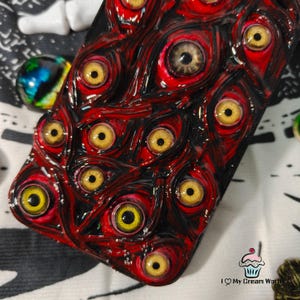 May include: A phone case with a unique design featuring numerous yellow-eyed, red-rimmed eyeballs. The case has a glossy finish and a dark background, creating a striking and unusual aesthetic. The design is intricate and detailed.