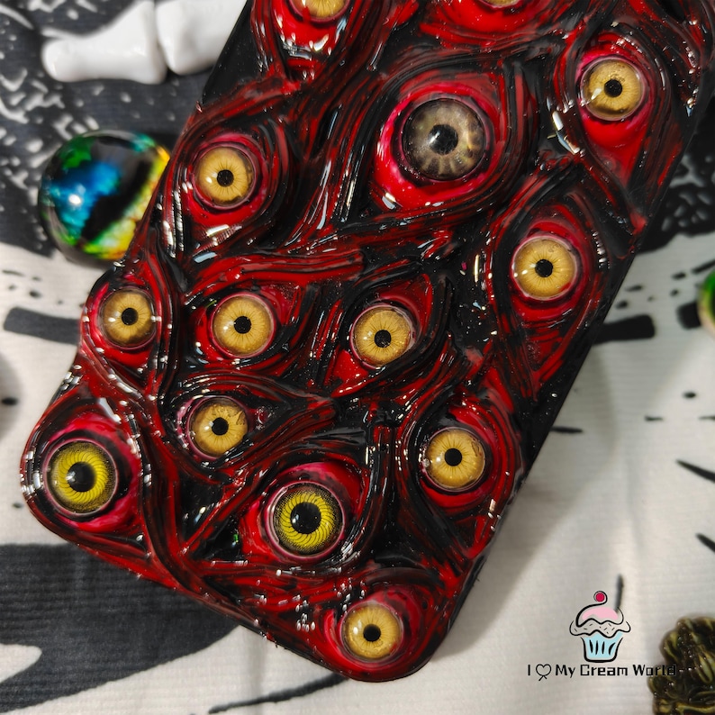 May include: A phone case with a unique design featuring numerous yellow-eyed, red-rimmed eyeballs. The case has a glossy finish and a dark background, creating a striking and unusual aesthetic. The design is intricate and detailed.