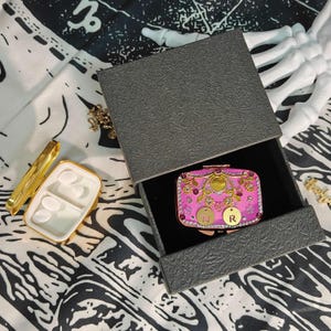 Peut inclure: Une boîte à bijoux rose et dorée décorative avec des ornements floraux et des pierres précieuses, présentée dans une boîte grise texturée. La boîte contient les lettres "N" et "R". Une petite boîte à pilules dorée est ouverte à côté.