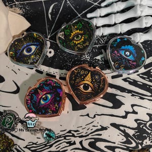 May include: Heart-shaped compact mirrors with eye designs. The mirrors feature various color schemes, including blue, green, and gold, with floral and celestial accents. Some mirrors have a rose gold frame. The overall theme is mystical and gothic.