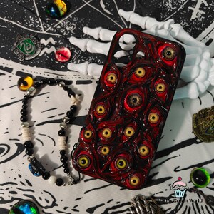 May include: A red and black phone case covered in multiple yellow-eyed, red-rimmed eyeballs. A black beaded bracelet with white and silver accents is next to the case. The background includes a white skeleton hand and other decorative items.