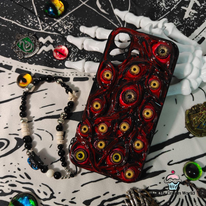 May include: A red and black phone case covered in multiple yellow-eyed, red-rimmed eyeballs. A black beaded bracelet with white and silver accents is next to the case. The background includes a white skeleton hand and other decorative items.