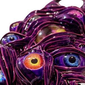 May include: A close-up of a purple, textured, 3D sculpture with multiple eyes. The eyes are painted in various colors, including blue, orange, and yellow.