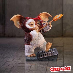 May include: A Gremlins figurine of a brown and white creature wearing a red headband and glasses, holding a torch. The figurine stands on a metal grate base. The Gremlins logo is in the bottom right corner.
