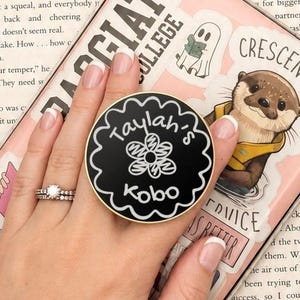 Könnte beinhalten: Ein schwarzer, runder Handyhalter mit gewelltem Goldrand, der den weißen Schriftzug Taylah's Kobo und ein handgezeichnetes Blumensymbol zeigt. Der Halter ist an einer hellrosa E-Reader-Hülle befestigt, die mit verschiedenen Stickern verziert ist.