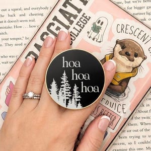May include: A circular black phone grip featuring the white text "hoa hoa hoa" above a graphic of pine trees. The grip is attached to a light pink phone case decorated with various stickers, including one of an otter and text reading "Crescent City".