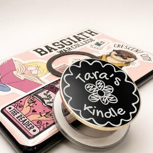 May include: A black circular phone grip with a scalloped edge, featuring a white flower illustration and the text "Tara's Kindle" in white. It is attached to a device case decorated with various stickers, including "Basgiath War College".