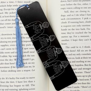 Peut inclure: Marque-page noir avec un pompon bleu clair. Le marque-page présente une illustration en contour blanc de quatre personnages portant des maillots avec les numéros 44, 22, 66 et 46. Le marque-page est posé sur un livre ouvert.