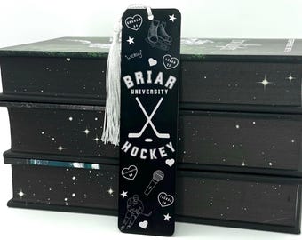 Op de afbeelding: Zwarte boekenlegger met een witte kwast, met de tekst "Briar University Hockey" en hockey-afbeeldingen. De boekenlegger is geplaatst voor een stapel zwarte boeken met een sterrenpatroon.