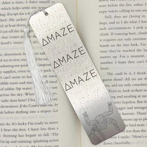 May include: Silver metal bookmark with the word "AMAZE" repeated three times. A small illustration of a figure is at the bottom. A white tassel hangs from the top.