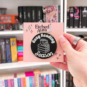 May include: A round black enamel pin with gold-toned edges attached to a light pink backing card. The pin features a white graphic of a stack of three books topped with a steaming mug and a leaf, with the text Cozy Reading Season in white.