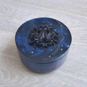 May include: A round, blue decorative box with a black octopus design on the lid. The box has a glossy finish and a dark blue color with lighter blue accents. The octopus is raised and detailed.
