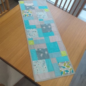 May include: A patchwork table runner with a geometric design in shades of grey, blue, and green. The runner is made of fabric squares with different patterns, including floral, leaf, and geometric designs.