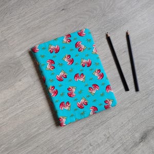 May include: A turquoise notebook featuring a pattern of red mushrooms and green leaves. Two black pencils lie beside the notebook on a grey wooden surface. The notebook is closed.