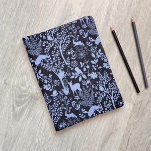 May include: A rectangular notebook with a black cover featuring a light blue pattern of trees, flowers, rabbits, and deer. Two black pencils are positioned diagonally to the right of the notebook, which is placed on a light wood grain surface.