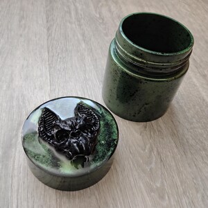 May include: A cylindrical jar with a matching lid, both featuring a dark green and black mottled finish. The lid is adorned with a raised, three-dimensional black bat sculpture centred on its surface, creating a textured and detailed appearance.