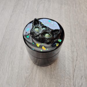 May include: A cylindrical black resin container featuring a three-eyed cat head sculpture on the lid. The lid surface is embedded with iridescent, multi-coloured foil flakes that shimmer in shades of green, blue, and yellow against the dark base.