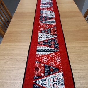 May include: A decorative table runner in red and black, featuring a central design of patterned triangles in white, navy, and red. The runner is placed on a light wood table, with a black trim and red border.