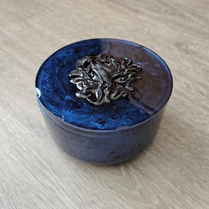 May include: A round, dark blue resin storage jar with a glossy, marbled finish. The lid features a prominent, raised, metallic black decorative emblem with an intricate, swirling, organic pattern centered on the top surface of the container.
