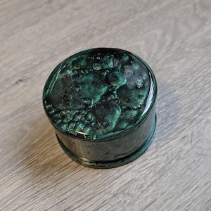 May include: A small, round, dark green box with a textured, glossy lid. The lid's surface has a mottled appearance, with darker and lighter shades of green. The box is sitting on a light gray wooden surface.