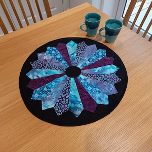 May include: A round, black table runner with a colourful, quilted design. The design features a circular pattern of blue, purple and white fabric pieces arranged in a radial pattern.