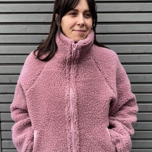 Könnte beinhalten: Eine altrosa Fleecejacke mit langen Ärmeln, einem hohen Kragen und einem durchgehenden Reißverschluss auf der Vorderseite. Die Jacke besteht aus einem strukturierten Sherpa-Material, hat Raglanärmel und zwei seitliche Eingrifftaschen.