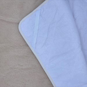 Puede incluir: Primer plano de una manta acolchada azul claro con borde blanco y superficie beige claro. La manta tiene un patrón de diamantes y un detalle de cinta azul claro. La manta está doblada, revelando el interior.