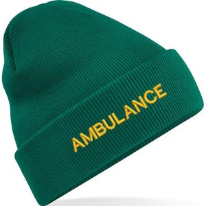 May include: A dark teal beanie hat with the word "AMBULANCE" embroidered in yellow. The hat is made of a ribbed knit fabric and has a folded brim. The text is centered on the brim.