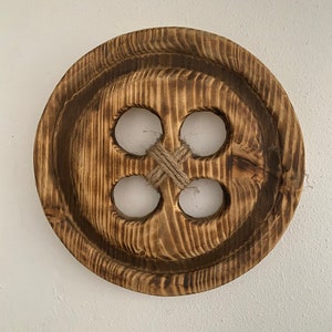 May include: A large, round, wooden button with four holes and a knotted twine detail. The button is made of natural wood with a dark brown stain.