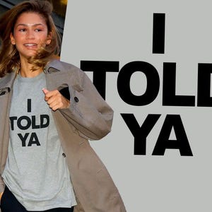 Puede incluir: Una mujer con una camiseta gris con el texto "I told ya" impreso en ella. Lleva un abrigo marrón y tiene el pelo largo y castaño. El fondo es una pared gris con el texto "I told ya" impreso en negro.