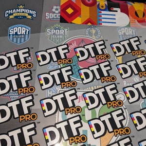May include: Close-up of multiple white stickers with the text "DTF PRO" in black letters. The stickers feature a small yellow, blue, and red flag design.