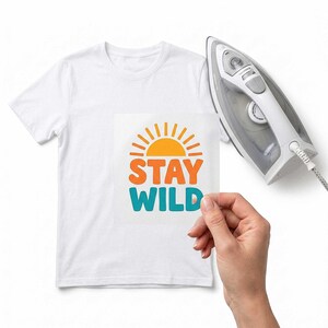Può includere: Una t-shirt bianca con una decalcomania a trasferimento termico raffigurante un sole arancione sopra il testo arancione STAY e il testo verde acqua WILD, posizionata accanto a un ferro da stiro elettrico argento e bianco per il tessuto.