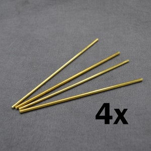 Könnte beinhalten: Vier goldfarbene Metallstäbe, jeder etwa 25 cm lang, sind fächerförmig auf einer grauen Oberfläche angeordnet. Der Text "4x" ist in Schwarz unter den Stäben gedruckt.