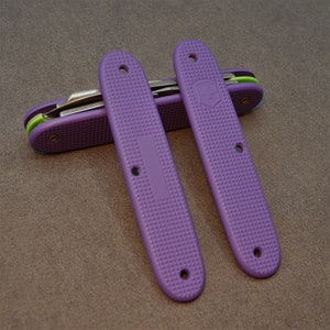 Könnte beinhalten: Zwei lilafarbene Taschenmesser-Schalen mit einer strukturierten Oberfläche. Die Schalen bestehen aus einem strapazierfähigen Kunststoff und haben ein charakteristisches Muster.