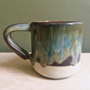 May include: A handcrafted ceramic mug with a distinctive glaze. The mug has a white base, a dark purple mid-section, and a top section with green, blue, and brown tones. The handle is green and brown. The mug is designed for hot drinks.