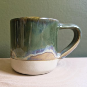 May include: A ceramic mug with a rounded body and a curved handle. The mug has a two-tone glaze: a creamy white base and a glossy green top with blue and gold accents. The mug is suitable for hot drinks.